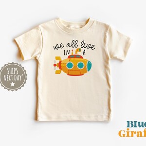 May include: A white t-shirt with a yellow submarine illustration and the text "we all live in a".
