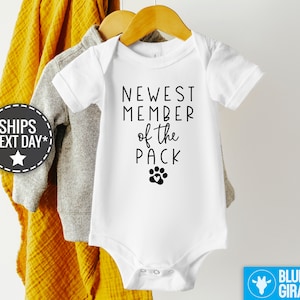May include: White baby onesie with black text that reads "Newest Member of the Pack" with a paw print below the text.