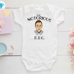May include: White baby onesie with a black graphic of Ruth Bader Ginsburg, the text "The Notorious R.B.G."