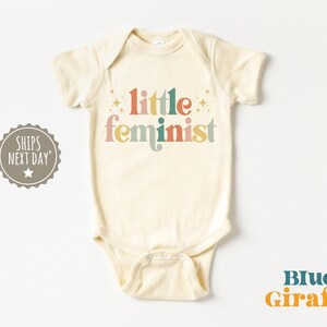May include: A cream coloured baby onesie with the text "little feminist" in colourful retro lettering with gold stars.