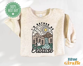May include: A beige sweatshirt with a graphic design of a mountain range, a waterfall, a tent, and a sun. The text "I'd rather be outside" is printed on the sweatshirt.
