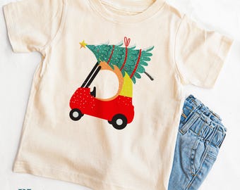 以下が含まれることがあります： クリーム色の半袖Tシャツ。赤いゴルフカートが、黄色い星を頂点に乗せた緑色のクリスマスツリーを運んでいるグラフィックが描かれています。カートには黒い車輪が付いています。Tシャツは青いデニムジーンズと組み合わされています。