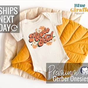 May include: White baby onesie with the text "Hippie Soul" in orange and pink lettering with floral accents. The onesie is on a yellow and beige textured surface.