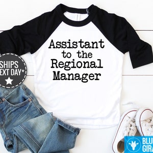 May include: A toddler's white and black baseball raglan shirt with the text 'Assistant to the Regional Manager' printed in a typewriter-style font. The shirt is displayed on a white surface with a pair of blue jeans and white canvas shoes. A small graphic shows the words 'SHIPS NEXT DAY'.