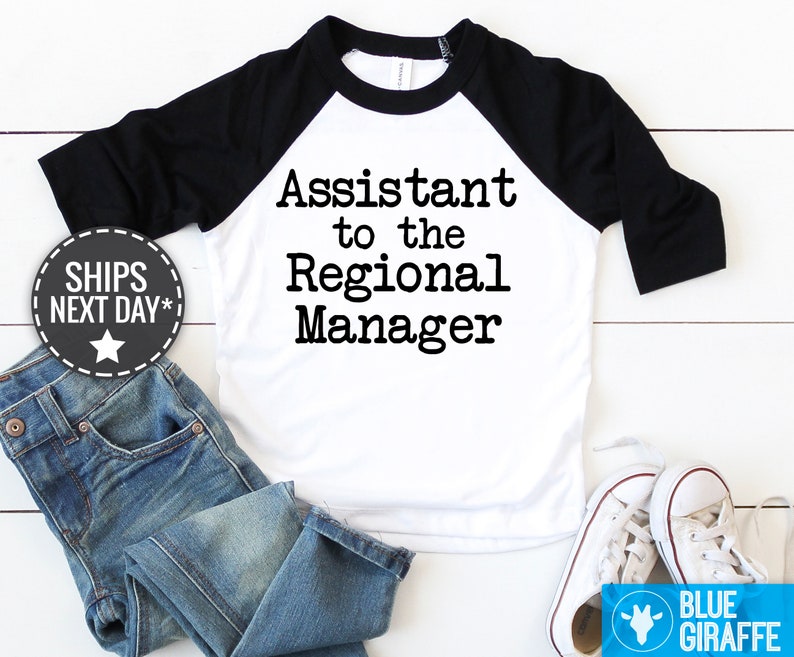 May include: A toddler's white and black baseball raglan shirt with the text 'Assistant to the Regional Manager' printed in a typewriter-style font. The shirt is displayed on a white surface with a pair of blue jeans and white canvas shoes. A small graphic shows the words 'SHIPS NEXT DAY'.