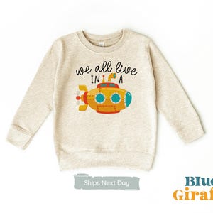 May include: A beige sweatshirt with the text "we all live in a" and a cartoon yellow submarine with blue portholes.