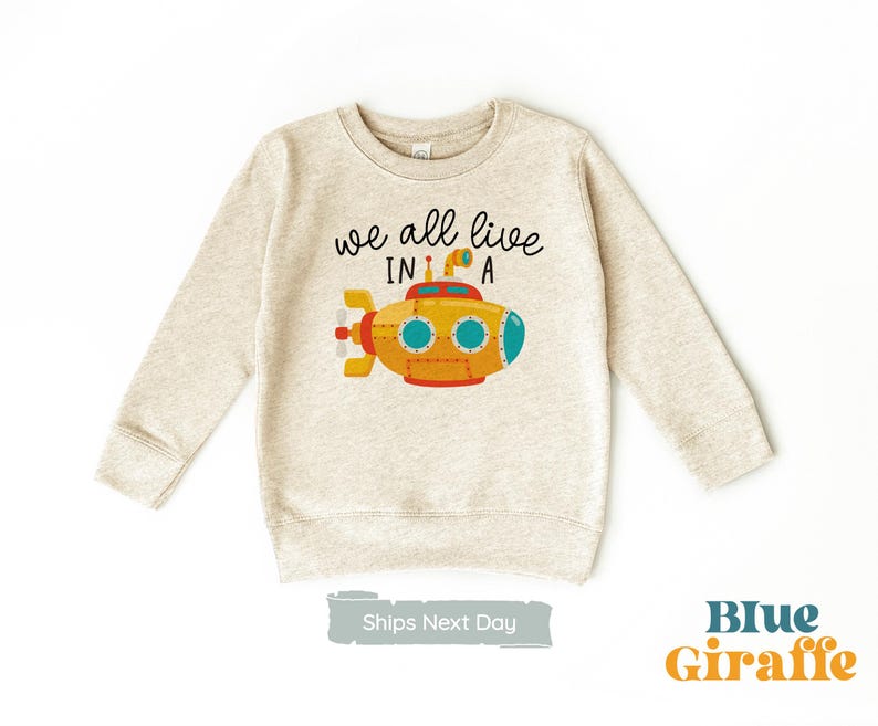 May include: A beige sweatshirt with the text "we all live in a" and a cartoon yellow submarine with blue portholes.