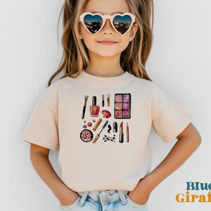 Pode incluir: Uma camiseta bege clara com uma ilustração em aquarela de itens de maquiagem, incluindo batom, sombra e pincéis. O texto "Blue Giraffe" é impresso em laranja e azul no canto inferior direito.