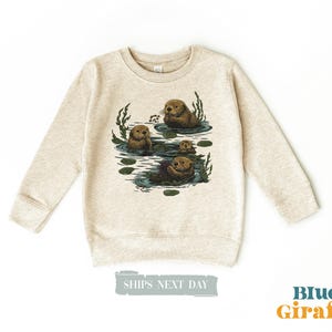 May include: Beige sweatshirt featuring a graphic of several otters in a pond with lily pads and aquatic plants. The otters are brown and the water is blue. The text "Blue Giraffe" is in orange and blue.
