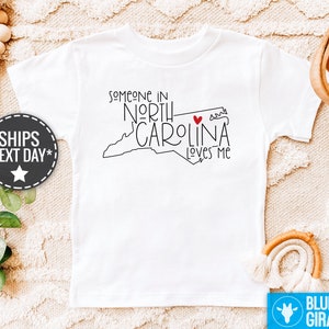 May include: White t-shirt with a black outline of the state of North Carolina and the text "Someone in North Carolina loves me" with a red heart.