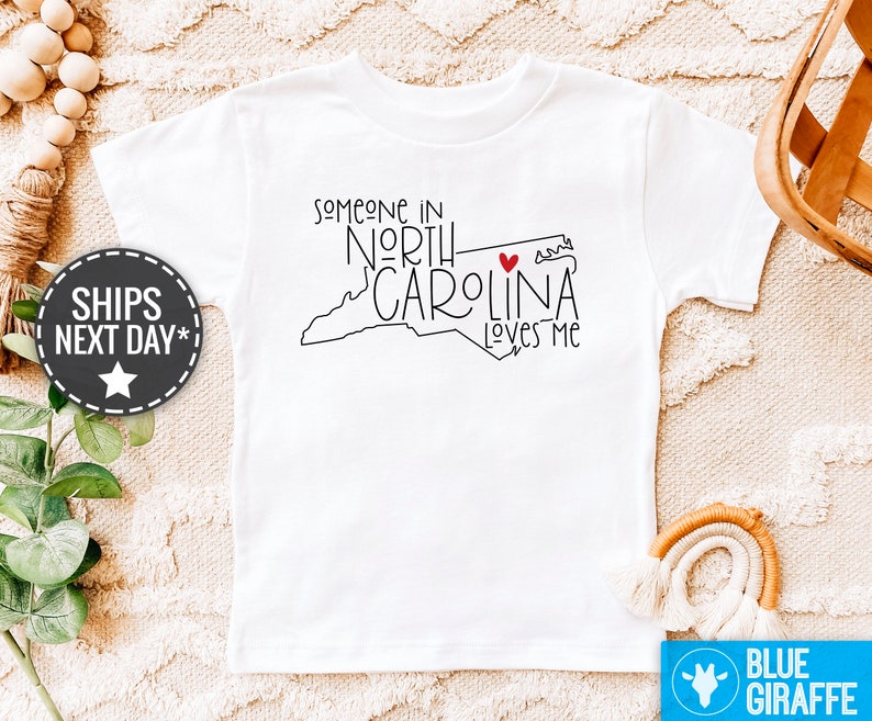 May include: White t-shirt with a black outline of the state of North Carolina and the text "Someone in North Carolina loves me" with a red heart.