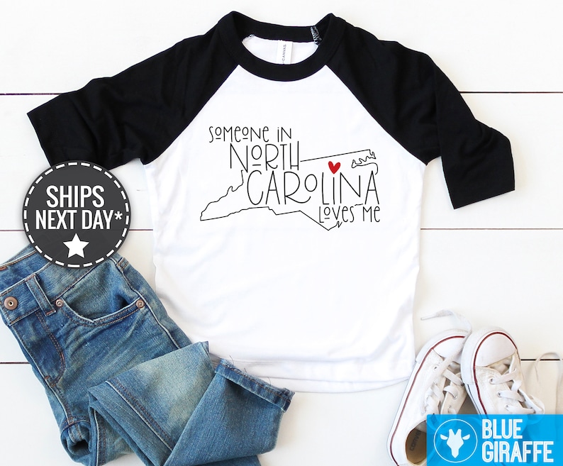 May include: White and black baseball tee shirt with a black outline of the state of North Carolina and the text "Someone in North Carolina loves me". A red heart is in the outline of the state.