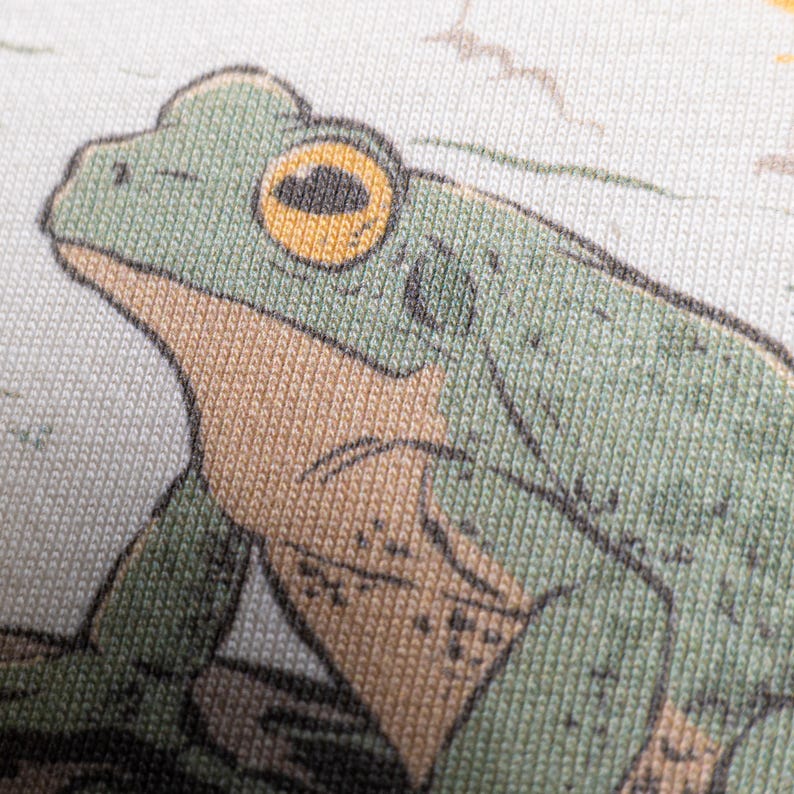 Puede incluir: Una vista de cerca de un estampado en tela que muestra una rana verde con el vientre beige y un ojo amarillo prominente. La ilustraci&oacute;n est&aacute; realizada con contornos oscuros sobre una superficie textil clara y texturizada con fibras visibles.