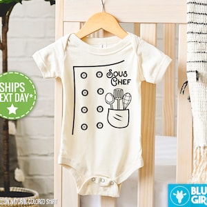 May include: A white baby onesie with a black design of a chef's uniform with buttons, a pocket, and the text "Sous Chef".  The pocket contains a spoon, a spatula, and a whisk.