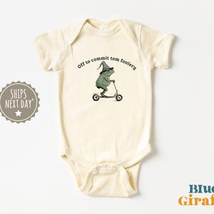 Op de afbeelding: Crèmekleurige baby-romper met een grillige kikkerillustratie. De kikker, met een tovenaars hoed, rijdt op een scooter. Tekst boven de kikker luidt "Off to commit tom foolery". Het Blue Giraffe-logo staat rechtsonder.