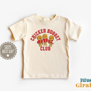 Pode incluir: Camiseta creme com a frase "CHICKEN NUGGET CLUB" em vermelho, com três desenhos animados de pepitas de frango vestindo jaquetas vermelhas. O logotipo Blue Giraffe está no canto inferior direito.