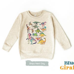 May include: A light beige sweatshirt featuring a colourful dinosaur print. The dinosaurs are all different sizes and shapes, and they are arranged in a random pattern. The sweatshirt has a crew neck and long sleeves.