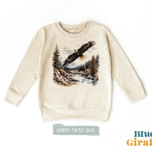 Puede incluir: Una sudadera beige con un estampado gráfico de un águila calva volando sobre una escena de bosque. El águila es marrón y blanca con un pico amarillo. El bosque es verde y marrón con un río azul que lo atraviesa.
