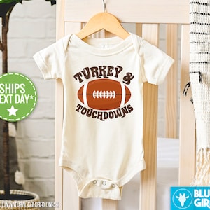 Op de afbeelding: Een witte baby romper met een bruine voetbal en de tekst "TURKEY & TOUCHDOWNS".