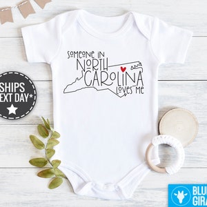May include: White baby onesie with black text that reads "Someone in North Carolina loves me". The text is accompanied by a black outline of the state of North Carolina with a red heart in the center.