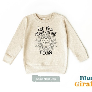 Puede incluir: Una sudadera beige con un gráfico negro de un dado de veinte caras y el texto "Let the Adventure Begin".