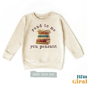 May include: A beige sweatshirt with the text "read to me you peasant" printed on the front. The graphic features a stack of books in various colors. The sweatshirt has long sleeves and a crew neck.