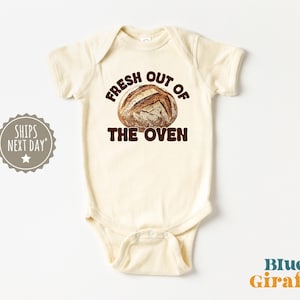 May include: Cream-colored baby bodysuit with the text "FRESH OUT OF THE OVEN" in brown letters and a graphic of a loaf of bread. The Blue Giraffe logo is in the bottom right corner.