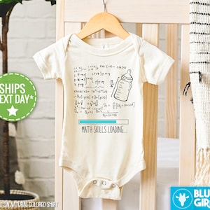 Könnte beinhalten: Ein weißer Baby-Strampler mit einer schwarz-weißen Illustration einer Babyflasche und mathematischen Gleichungen. Der Text "MATH SKILLS LOADING..." ist unter der Illustration gedruckt. Der Strampler hängt an einem Holzbügel.