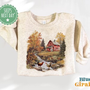 Könnte beinhalten: Ein beiger Pullover mit einer bunten Illustration einer roten Scheune, Bäumen und Hühnern. Die Illustration ist im Vintage-Stil gehalten.