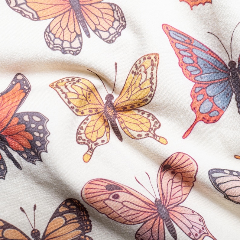 May include: A cream-colored fabric featuring a repeating pattern of butterflies in various shades of orange, yellow, blue, and brown. The butterflies are depicted with detailed wing veins and dark bodies, scattered across the textile surface.