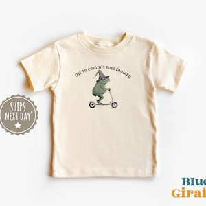 May include: A cream-coloured t-shirt with a humorous design. It shows a green frog in a wizard's hat riding a scooter, with the text "Off to commit tom foolery" above. The Blue Giraffe logo is visible. The shirt is a light, neutral colour.