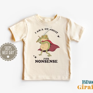 May include: Cream-colored t-shirt featuring a cartoon frog with a sword and cape. The frog is green with a red cape, holding a sword, and the text reads "I AM A CONDUIT FOR NONSENSE". The shirt has a round neck and short sleeves.