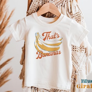 Puede incluir: Camiseta de color crema con un gráfico retro de plátanos y el texto "That's Bananas" en naranja y rojo. La camiseta está colgada en una percha de madera sobre un fondo neutro. Las palabras "Blue Giraffe" están en la esquina inferior derecha.
