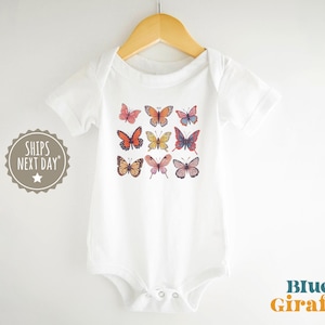 May include: A white short-sleeved baby bodysuit featuring a graphic print of nine colorful butterflies arranged in a grid pattern. The garment is displayed on a wooden hanger and includes a circular badge with the text SHIPS NEXT DAY.