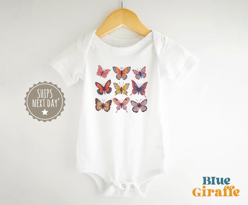 May include: A white short-sleeved baby bodysuit featuring a graphic print of nine colorful butterflies arranged in a grid pattern. The garment is displayed on a wooden hanger and includes a circular badge with the text SHIPS NEXT DAY.