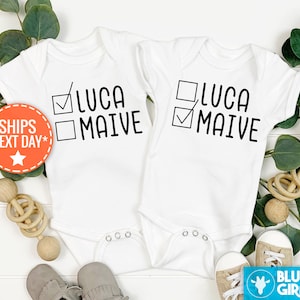 May include: Two white baby onesies with black text. One onesie says "LUCA" with a tick in the box next to it and "MAIVE" with an empty box next to it. The other onesie says "LUCA" with an empty box next to it and "MAIVE" with a tick in the box next to it.
