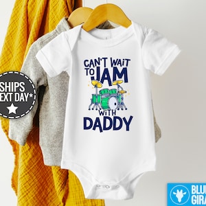 May include: White baby onesie with a blue and green graphic of a drum set and the text "Can't Wait to Jam with Daddy".