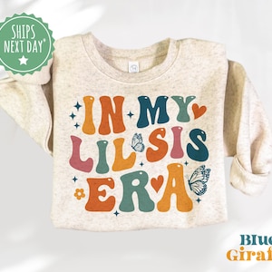 May include: A light beige sweatshirt with the text "In My Lil Sis Era" in colorful retro-style lettering. The text is decorated with butterflies and hearts.
