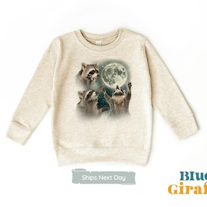 May include: A beige sweatshirt with a graphic print of three raccoons looking up at a full moon. The raccoons are in a playful pose, with one reaching up to touch the moon. The sweatshirt has a crew neck and long sleeves.
