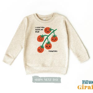 May include: A heather beige long-sleeve sweatshirt featuring a graphic of a vine with four red tomatoes, each with a different cartoon face. The text "I LOVE YOU FROM MY HEAD" is printed above the tomatoes, and "TOMATOES" is printed below them.
