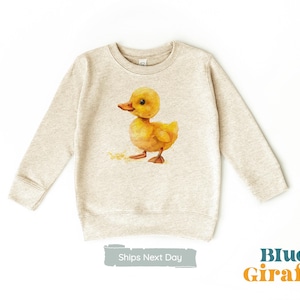 Może przedstawiać: Beżowa bluza z nadrukiem żółtego kaczątka. Kaczaczek jest zwrócony w prawo i ma żółty dziób oraz czarne oczy. Bluza ma długie rękawy i okrągły dekolt.