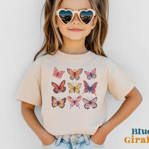 May include: A beige short-sleeve t-shirt featuring a graphic print of nine colorful butterflies in various patterns and shades of orange, pink, yellow, and blue. The shirt is paired with heart-shaped sunglasses and light blue denim jeans.