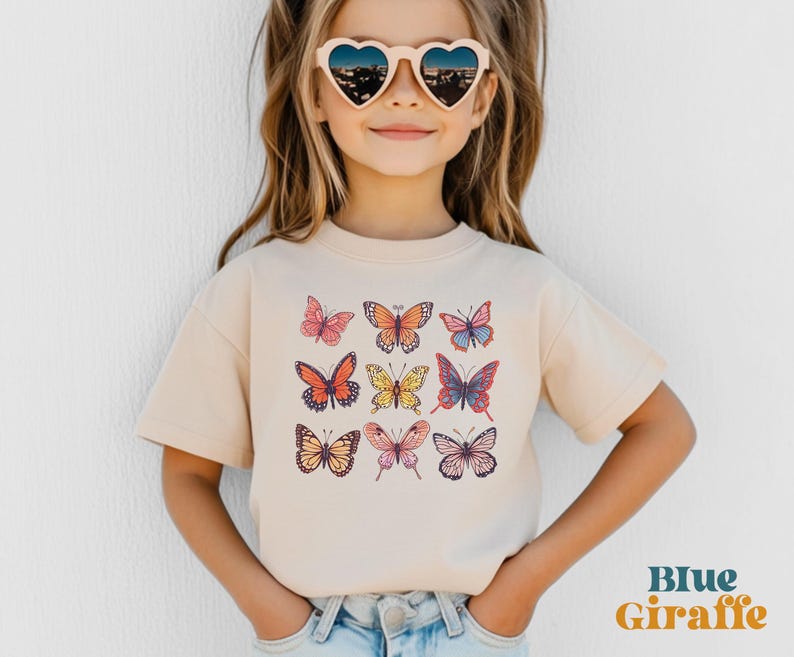 May include: A beige short-sleeve t-shirt featuring a graphic print of nine colorful butterflies in various patterns and shades of orange, pink, yellow, and blue. The shirt is paired with heart-shaped sunglasses and light blue denim jeans.