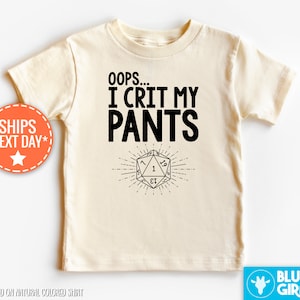 May include: A natural-colored short-sleeve t-shirt featuring the black text OOPS... I CRIT MY PANTS above a graphic of a twenty-sided die with the number one centered on the face, surrounded by radiating lines, printed on the front of the garment.