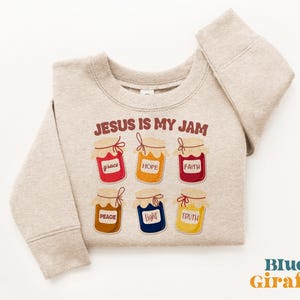 Könnte beinhalten: Beiges Sweatshirt mit dem Text "JESUS IS MY JAM" und Illustrationen von sechs Marmeladengläsern. Jedes Glas hat eine andere Farbe und Aufschrift: Gnade, Hoffnung, Glaube, Frieden, Licht und Wahrheit. Das Blue Giraffe-Logo ist unten rechts zu sehen.