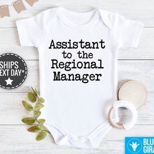 May include: White baby bodysuit with black text reading, 'Assistant to the Regional Manager'.  The bodysuit is shown on a white wooden surface with greenery and a wooden baby teether.  The words 'SHIPS NEXT DAY*' are on a dark gray star-shaped label. A light blue label with the words 'BLUE GIRAFFE' is in the bottom right corner.