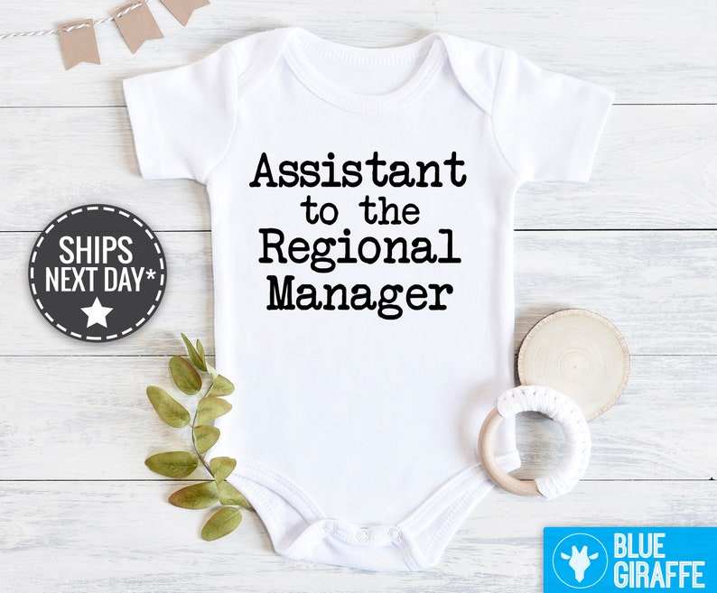 May include: White baby bodysuit with black text reading, 'Assistant to the Regional Manager'.  The bodysuit is shown on a white wooden surface with greenery and a wooden baby teether.  The words 'SHIPS NEXT DAY*' are on a dark gray star-shaped label. A light blue label with the words 'BLUE GIRAFFE' is in the bottom right corner.