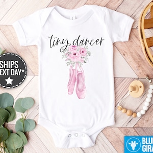 May include: White baby bodysuit with the text "tiny dancer" in black script. Pink watercolor flowers and ballet shoes are pictured below the text.