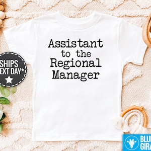 May include: White toddler t-shirt featuring black text that reads, 'Assistant to the Regional Manager.' The shirt is shown on a light beige surface with decorative elements.  The phrase is a reference to popular culture. The image also includes a small graphic indicating next-day shipping and a watermark with the words 'Blue Giraffe'.