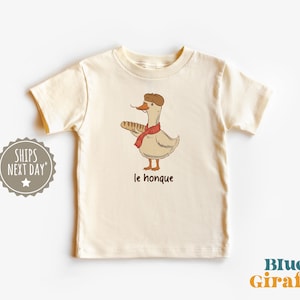 Puede incluir: Camiseta color crema con un dibujo de un ganso de dibujos animados con boina y bufanda, sosteniendo una baguette. El texto "le honque" está debajo. La camiseta tiene mangas cortas y cuello redondo. El logo de Blue Giraffe está abajo a la derecha.